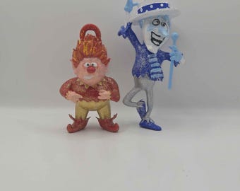 Puede incluir: Dos figuras brillantes: una es un personaje rojo y dorado con pelo puntiagudo y un lazo en la cabeza, y la otra es un personaje azul y blanco con sombrero, que sostiene un bastón y está de pie sobre una pierna con un lazo en el sombrero.