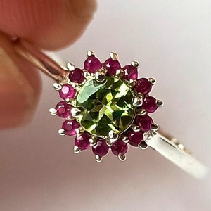 May include: A silver ring with a green gemstone surrounded by a halo of red gemstones.