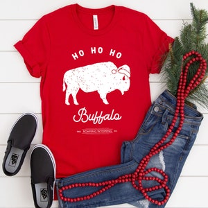 May include: Red t-shirt with a white buffalo wearing a Santa hat. The text "Ho Ho Ho Buffalo" is printed on the shirt. The text "The Roaming Wyoming Co." is printed below the buffalo.