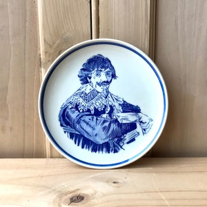Peut inclure: Une assiette en céramique blanche avec une illustration bleue et blanche d'un homme avec une moustache et un grand col. L'assiette a un bord bleu.
