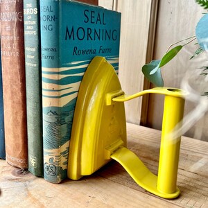 May include: A vibrant yellow, retro-style iron bookend holding up a collection of old books. The bookend is designed to resemble an iron, complete with a handle. The books include titles such as "Seal Morning" and "The Birds of Britain and Europe."