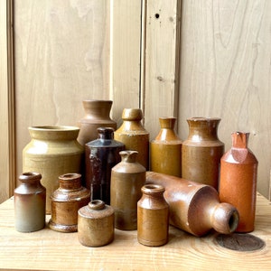 Puede incluir: Una colección de 12 botellas de cerámica de gres marrón vintage de varios tamaños y formas. Las botellas están dispuestas sobre una superficie de madera.