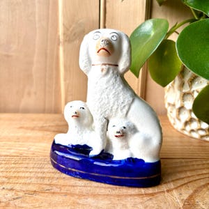 Puede incluir: Figura de perro de cerámica blanca con dos cachorros, sobre una base azul con ribete dorado. El perro tiene un collar marrón y detalles rojos. La figura está sobre una superficie de madera.
