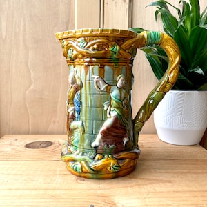 May include: A ceramic pitcher with a brown, green, and yellow glaze. The pitcher features a raised design of people and a building. The handle is shaped like a twisted vine.