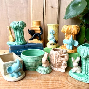 May include: A selection of vintage ceramic vases and planters in various shapes and colours. The collection includes a green palm tree vase with a giraffe, a black cat on a yellow base, and a yellow vase with a poodle. Other planters feature elephants, rabbits, and monkeys.