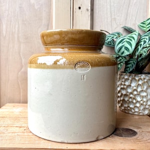 May include: A cylindrical stoneware crock featuring a two-tone design with a glossy brown glazed rim and a matte cream-coloured base. The front displays an impressed circular stamp reading Caledonian Rutherglen and the number 16 below the mark.