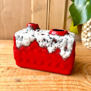 Peut inclure: Un vase en céramique rouge vif avec un dessus texturé blanc et noir. Le vase a deux ouvertures et une forme rectangulaire. Le vase est posé sur une surface en bois.