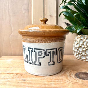 May include: A vintage ceramic storage jar with a lid, displaying the word "LIPTON" in black lettering. The jar has a cream-coloured base and a brown lid and rim. The jar is likely for storing tea or other dry goods.