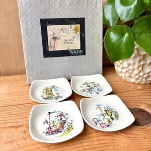 May include: A set of four vintage Wade of England ceramic dishes. Each square dish features a colourful, whimsical illustration. The dishes are white with a slightly curved edge. A grey box with the text "EMETTS ORIGINAL POTTERY" is in the background.