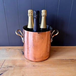 May include: A cylindrical copper wine cooler featuring two polished brass side handles. The vessel contains three glass bottles, two with gold foil-wrapped necks and one with a black foil-wrapped neck, positioned upright inside the metallic container.