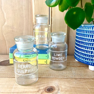 May include: Three clear glass jars with white labels and white lettering. The labels read "Borax", "Lime Powder", and "Ferrous Sulphate". The jars are sitting on a wooden surface with a stack of books behind them.