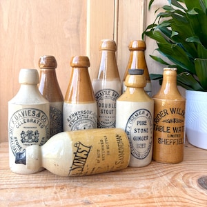 Puede incluir: Una colección de botellas antiguas de gres en varias formas y tamaños. Las botellas presentan etiquetas con texto y logotipos, en tonos beige, marrón y crema. Algunas botellas están de pie, mientras que una está horizontal.