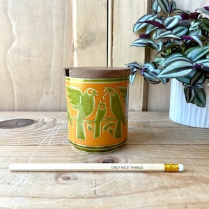 May include: A ceramic jar with a wooden lid, featuring a design of green birds on an orange background. The jar is cylindrical and sits on a wooden surface. A pencil with the text "ONLY NICE THINGS" is also present.