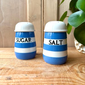 May include: A pair of ceramic salt and sugar shakers. The shakers are blue with white stripes and white lids. The word "SUGAR" is printed on one shaker, and "SALT" is printed on the other. The shakers are on a wooden surface.
