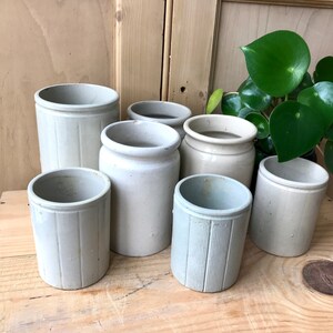 May include: Seven white ceramic jars with a ribbed design. The jars are of various sizes and are arranged in a row on a wooden surface.