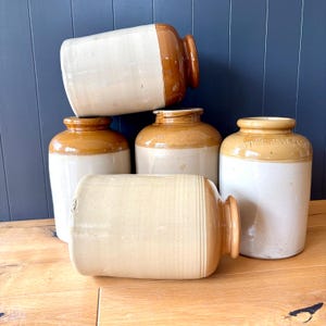 May include: A group of vintage stoneware jars in different shapes and sizes. The jars are mainly white with brown detailing, including rounded and rectangular designs. One jar is inscribed with the text "STOCKMAST PASTE".