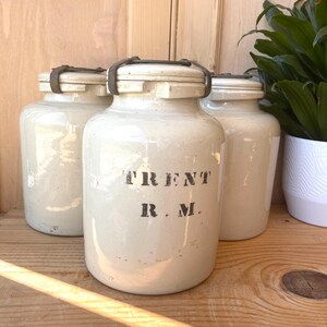 May include: Three vintage white ceramic jars with metal lids. The middle jar has the text "TRENT R.M." printed on the front.
