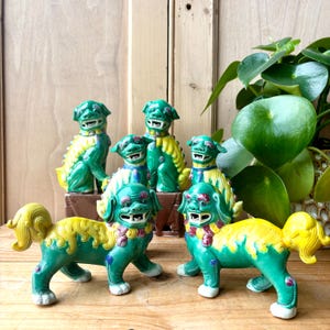 Puede incluir: Una colección de estatuas de perros Foo de cerámica verde y amarilla. Las estatuas tienen detalles, incluyendo una expresión feroz, detalles ornamentados y una paleta de colores vibrantes. Los perros Foo están dispuestos sobre una superficie de madera, con una planta en el fondo.