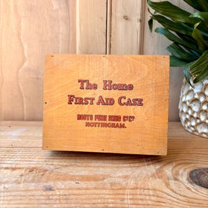 Peut inclure: Un ancien coffret de premiers secours en bois avec le texte "The Home FIRST AID CASE" en lettres rouges et noires. Le coffret porte également le texte "BOOTS PURE DRUG C. LTD. NOTTINGHAM." Le coffret est posé sur une surface en bois.