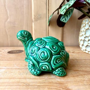 Op de afbeelding: Een glanzend, smaragdgroen keramieken schildpadbeeldje met een verhoogd, wervelend schildpatroon en een getextureerde huid op de poten. De schildpad is afgebeeld in zijaanzicht met de kop iets omhoog gedraaid, wat een gladde, reflecterende afwerking laat zien.