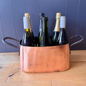 May include: An oval copper container with two dark metal side handles, holding five glass wine bottles. The copper vessel has a smooth, reflective surface with visible hammered texture and rounded corners, containing bottles with various coloured caps.