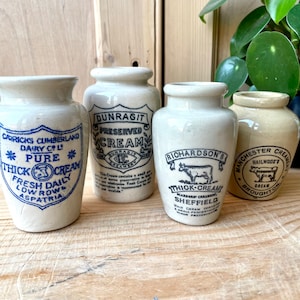 Peut inclure: Quatre pots à crème anciens avec des étiquettes bleues et noires. Les pots portent diverses inscriptions, notamment "Thick Cream" et des noms de marques comme "Richardson's". Les pots sont de couleur blanc cassé et ont une esthétique vintage.