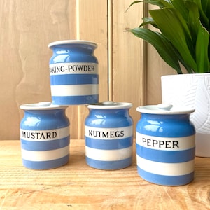 Peut inclure: Quatre pots à épices en céramique à rayures bleues et blanches, étiquetés "BAKING-POWDER", "MUSTARD", "NUTMEGS" et "PEPPER". Les pots ont des couvercles blancs et sont disposés sur une surface en bois.