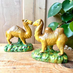 Könnte beinhalten: Zwei Kamel-Figuren aus Keramik. Die Kamele sind goldgelb mit braunen Akzenten und stehen auf grünen Sockeln. Die Figuren sind so positioniert, als würden sie miteinander interagieren. Der Hintergrund ist eine hellbraune Holzoberfläche.