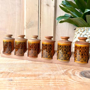 May include: A collection of six vintage spice jars with wooden lids and a wooden rack. The jars are a warm yellow-brown colour with black decorative patterns and labels. The labels include the names of spices.