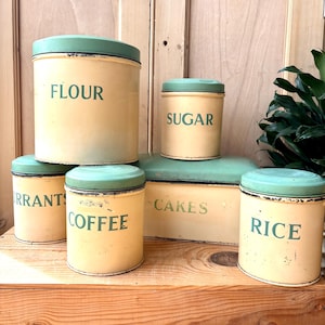 May include: A collection of vintage kitchen canisters and a cake tin. The canisters are cream-colored with green lids and feature labels for flour, sugar, currants, coffee, and rice. The cake tin is rectangular with a green lid and the word "CAKES".