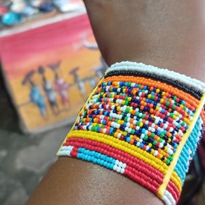 May include: A wide, beaded bracelet in a vibrant array of colors, including red, yellow, blue, and white. The bracelet is made of small, colorful beads and is worn on the wrist. The design features a mix of patterns and colors.
