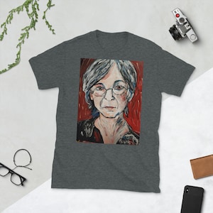 Könnte beinhalten: Ein graues T-Shirt mit einem farbenfrohen Porträt einer Person mit grauem Haar und Brille. Das Porträt ist in einer stilisierten Art mit roten, gelben und blauen Akzenten gemalt. Der Hintergrund ist ein rotes und oranges abstraktes Design.