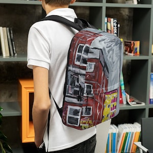 Puede incluir: Una mochila negra con correas ajustables presenta una impresión colorida de un edificio con acentos rojos, amarillos y grises. La impresión tiene un estilo pintado a mano.