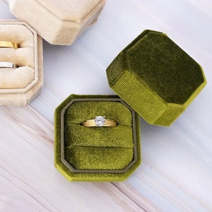 May include: A selection of velvet ring boxes in gold, cream, and olive green. The octagonal boxes are open, showcasing rings with diamonds and gold bands. The background is a light, marbled surface.