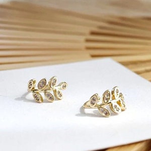 May include: A pair of gold-toned ear cuffs shaped like small leafy branches. Each piece features a series of marquise-shaped leaves, with each leaf inlaid with a small, clear, sparkling stone. The jewelry is displayed on a white card.