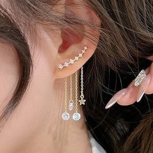 Può includere: Orecchino a cerchio dorato con una fila di piccole pietre di zircone cubico e catene pendenti con ciondoli a forma di stella e zircone cubico rotondo.