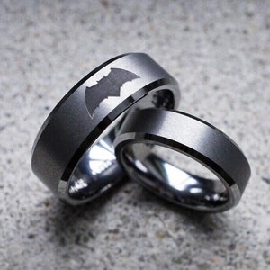 Peut inclure: Deux alliances en carbure de tungstène gris avec des bords biseautés. Une alliance présente un logo Batman noir.