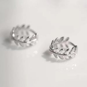 May include: Two silver-colored hoop earrings. Each earring features a leaf design around the hoop. The earrings are displayed on a white surface, with soft lighting and shadows.