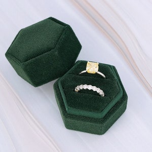 May include: Two dark green velvet ring boxes, one open, displaying two rings. One ring has a square yellow gemstone, the other has a row of small, clear stones. The boxes are hexagonal and sit on a white, textured surface.