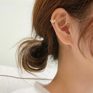 May include: A gold-toned ear cuff featuring a delicate leaf-inspired design with small, clear, sparkling stones embedded along the metal surface. The piece is positioned on the upper cartilage of the ear, showcasing a curved, branch-like structure.
