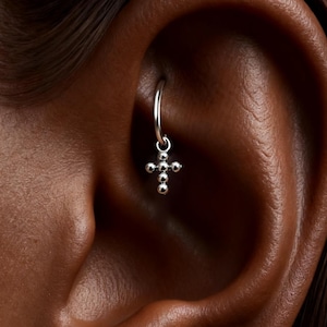 May include: A silver hoop earring with a small cross pendant. The cross is made of small, round beads. The earring is shown in a close-up view, highlighting the details of the jewelry.