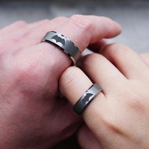 May include: Two silver rings with a black bat symbol engraved on each. The rings are worn on the fingers of two hands.