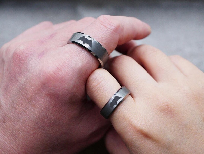 May include: Two silver rings with a black bat symbol engraved on each. The rings are worn on the fingers of two hands.