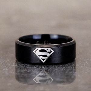 May include: A black tungsten carbide ring with a brushed finish and a silver Superman logo engraved on the top.