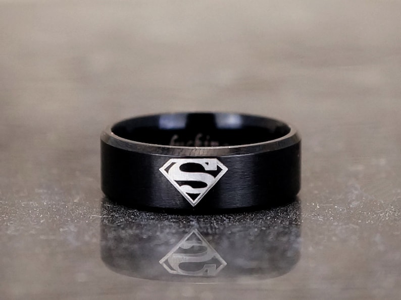 May include: A black tungsten carbide ring with a brushed finish and a silver Superman logo engraved on the top.