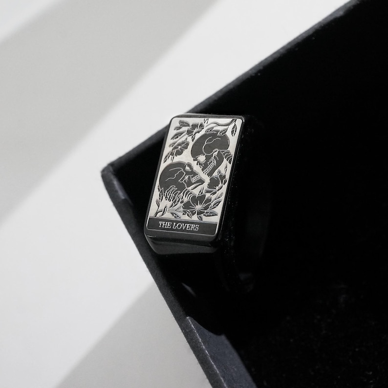May include: A black rectangular signet ring featuring a silver-toned engraved design of two skulls facing each other surrounded by floral motifs. The top of the ring displays the Roman numeral VI and the text THE LOVERS at the bottom edge.