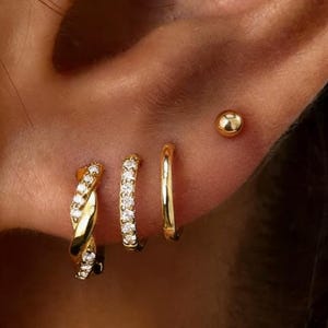 Könnte beinhalten: Nahaufnahme eines Ohrs mit goldenen Ohrringen. Das Set umfasst einen kleinen goldenen Kugelstecker, einen kleinen goldenen Reifen, einen goldenen Reifen mit klaren Steinen und einen verdrehten goldenen Reifen mit klaren Steinen.