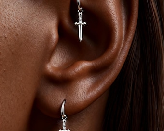 May include: Silver hoop earrings with a small sword pendant. The earrings are attached to the ear. The sword pendants have a detailed hilt and a pointed blade. The earrings are a simple, yet elegant design.