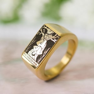 May include: A gold-toned signet ring featuring a rectangular face with a black and gold engraved illustration of a figure suspended upside down. The engraving includes the text THE HANGED MAN at the bottom of the rectangular face of the ring.