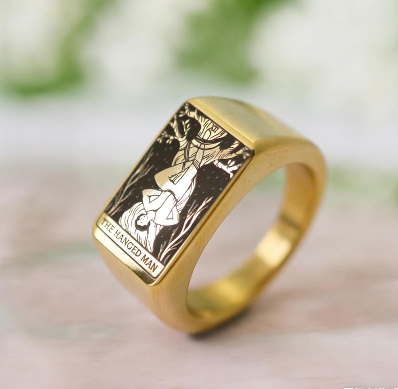 May include: A gold-toned signet ring featuring a rectangular face with a black and gold engraved illustration of a figure suspended upside down. The engraving includes the text THE HANGED MAN at the bottom of the rectangular face of the ring.